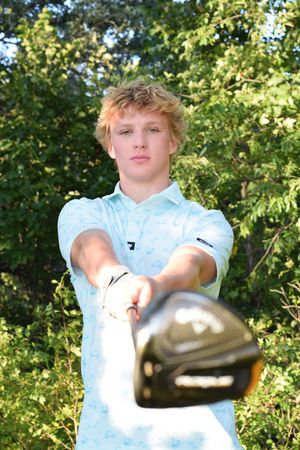 Golfer holding club towards camera with trees in background.