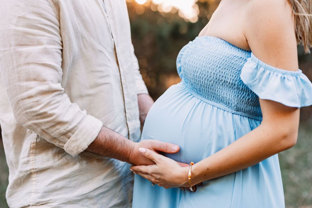 Close-up of hands tenderly touching pregnant belly in light blue off-shoulder dress.