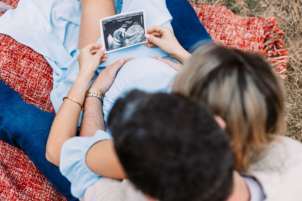 Couple looking at ultrasound photo while lying on red blanket outdoors.
