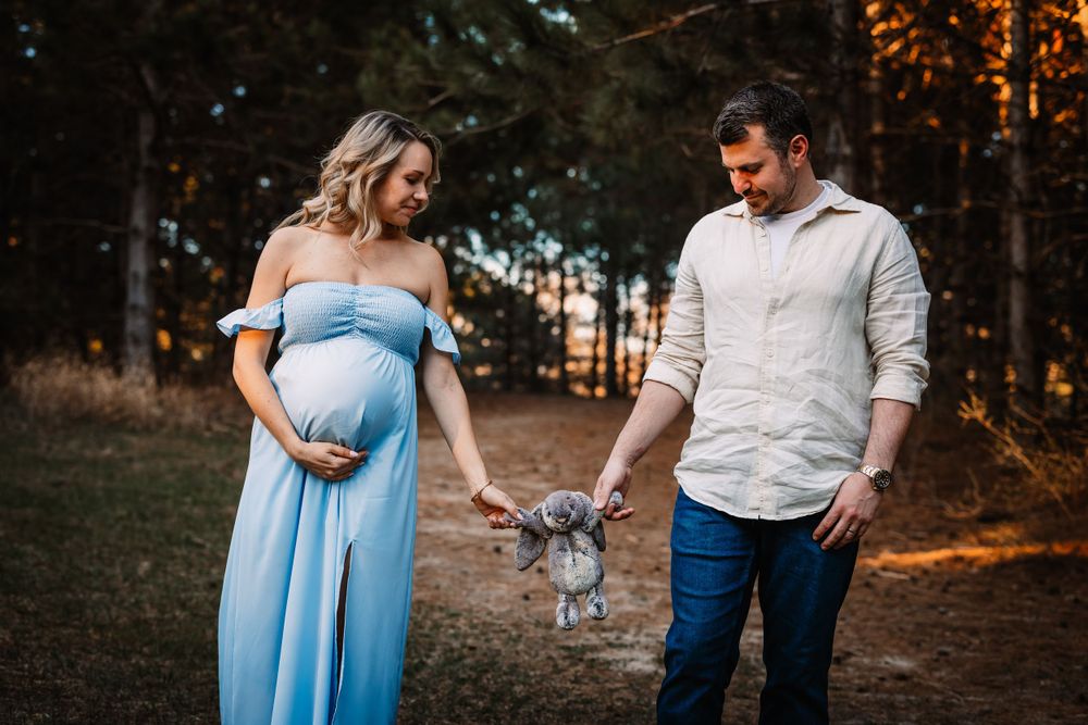 Maternity photoshoot in forest with couple holding baby clothes at golden hour.