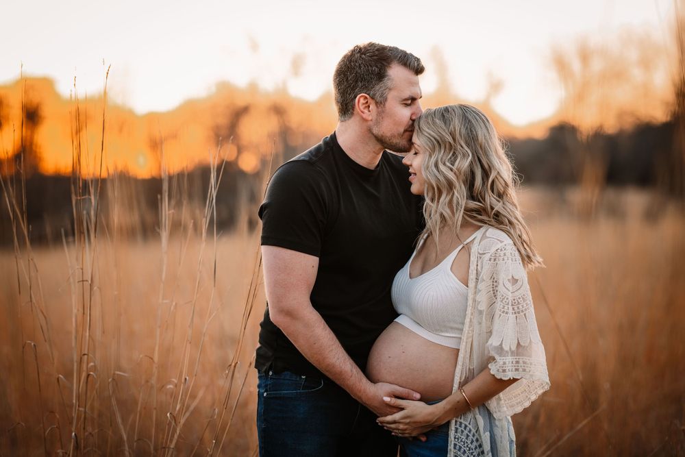 Intimate maternity photo of couple embracing in field at sunset with warm lighting.
