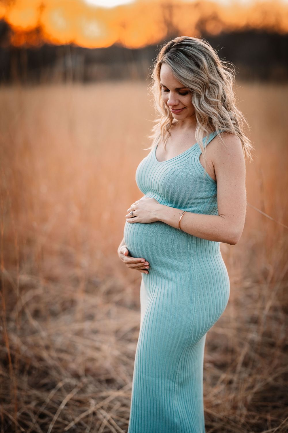 Pregnant woman in turquoise dress poses during golden sunset in field of tall grass.
