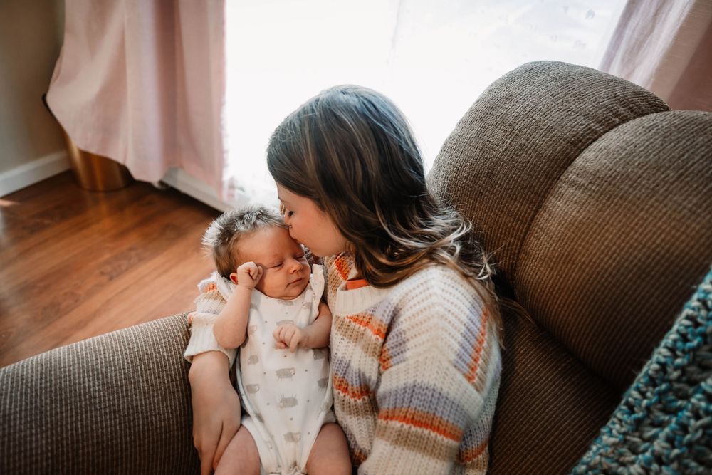 A tender moment between parent and newborn baby as they cuddle on a brown couch in a sunlit room.