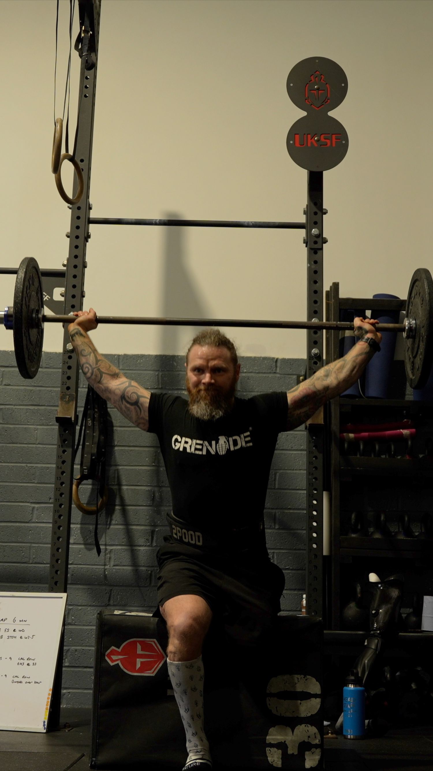 Weightlifter in Grenade shirt performing overhead press with barbell in gym setting.