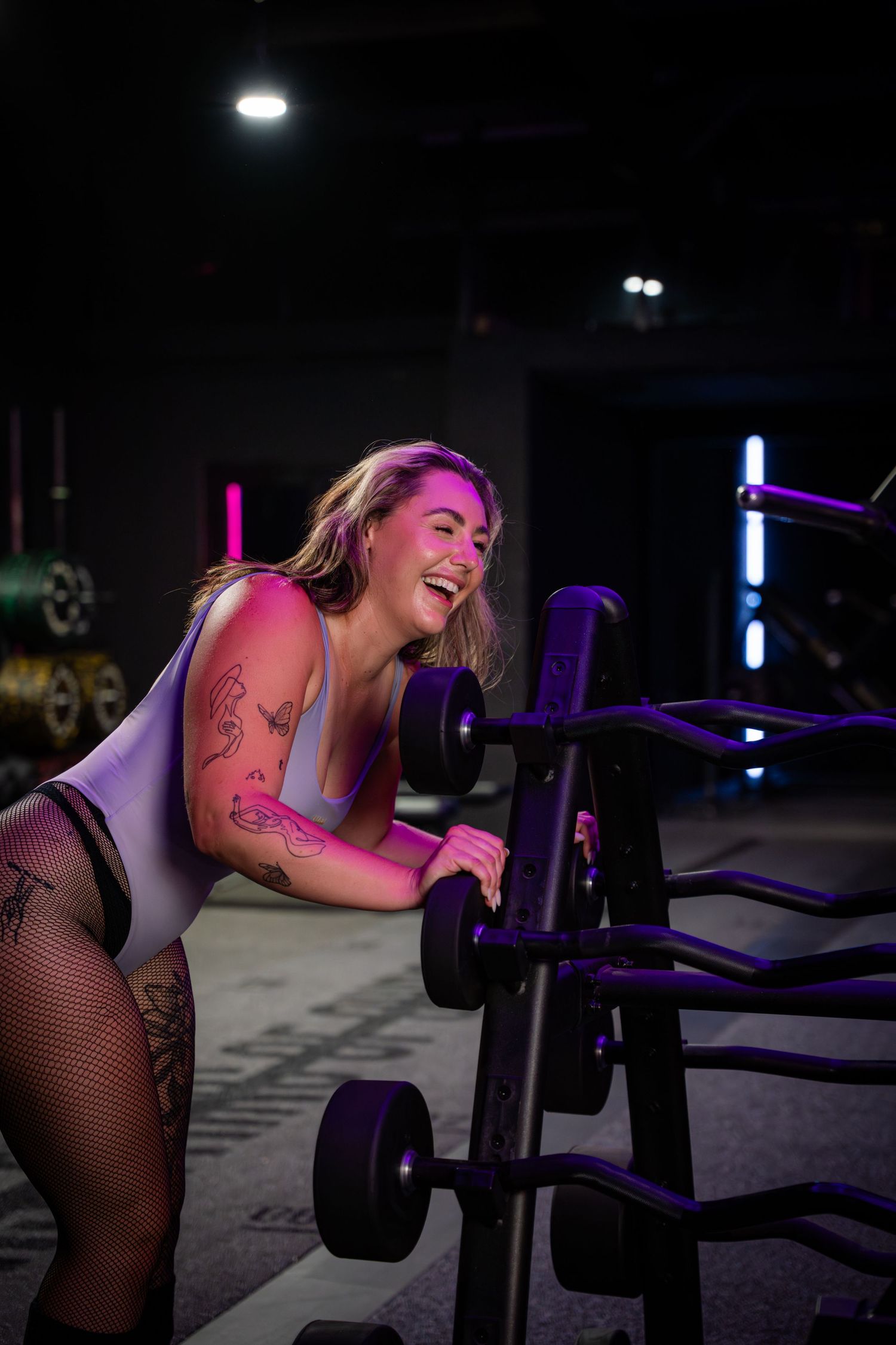 Fitness enthusiast laughing while working out with weights in neon-lit gym setting.