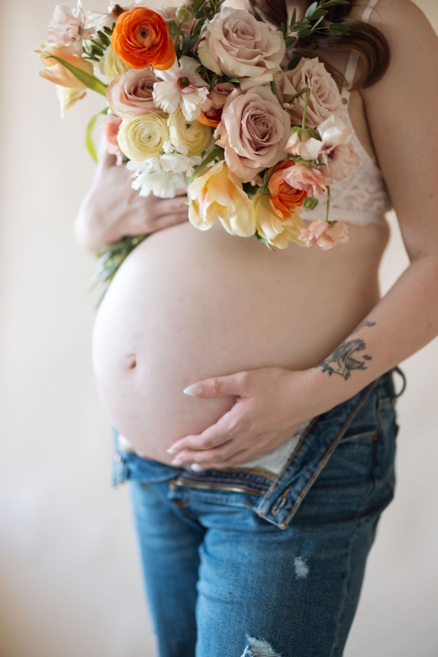 A colorful bouquet of roses rests against a pregnant belly in jeans.