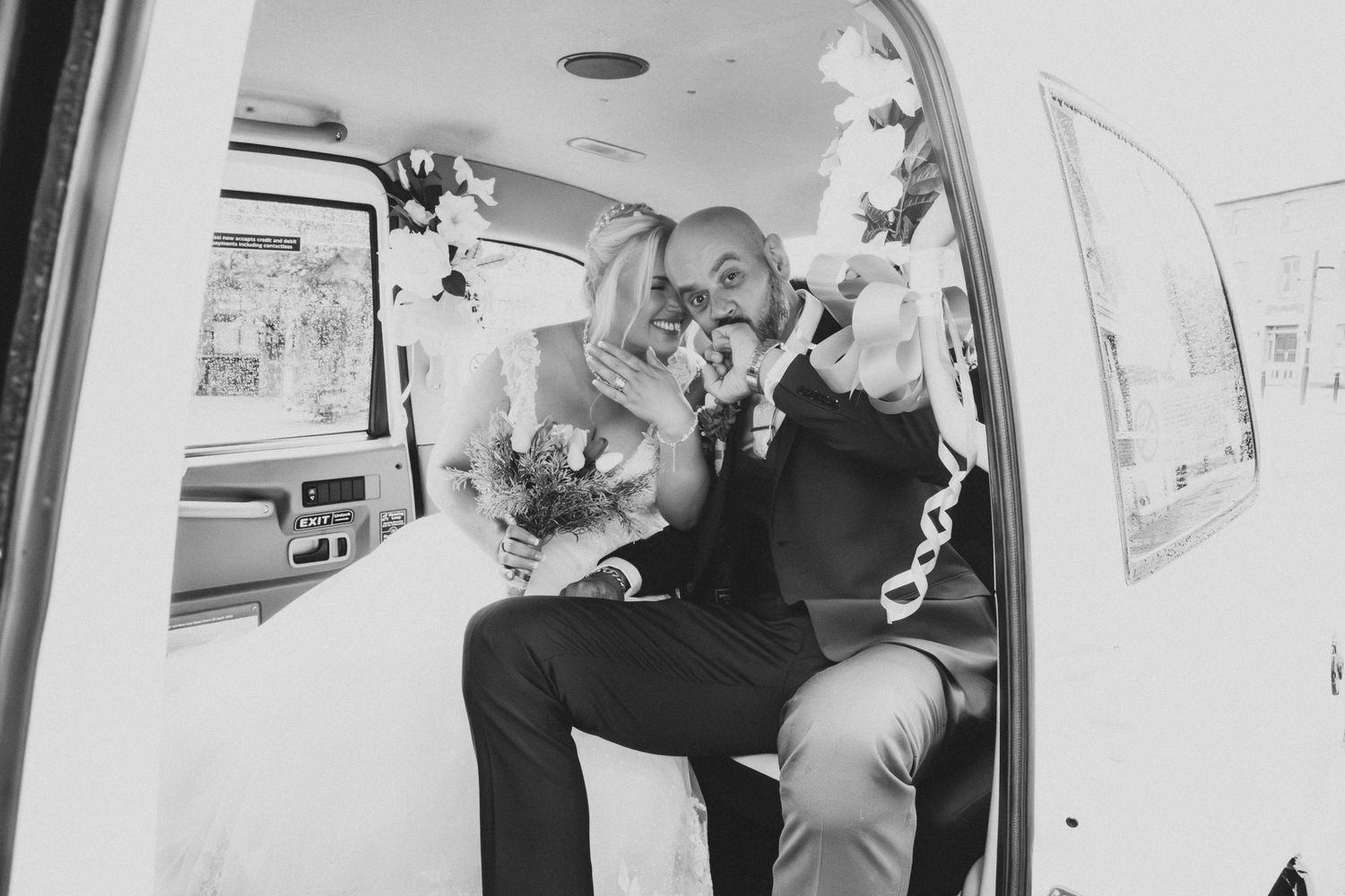 Black and white photo of a bride and groom sitting in a decorated car, sharing a joyful moment on their wedding day.