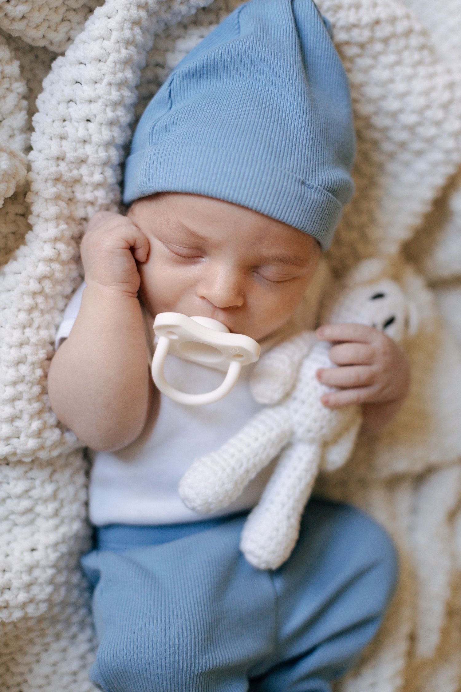 Peaceful sleeping baby in blue hat with pacifier and white knit stuffed animal on cozy blanket.