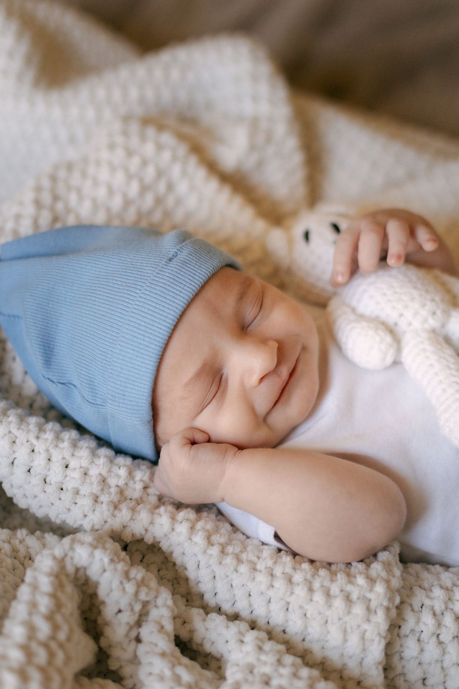 Peaceful sleeping newborn wrapped in white knit blanket wearing soft blue cap and holding plush toy.