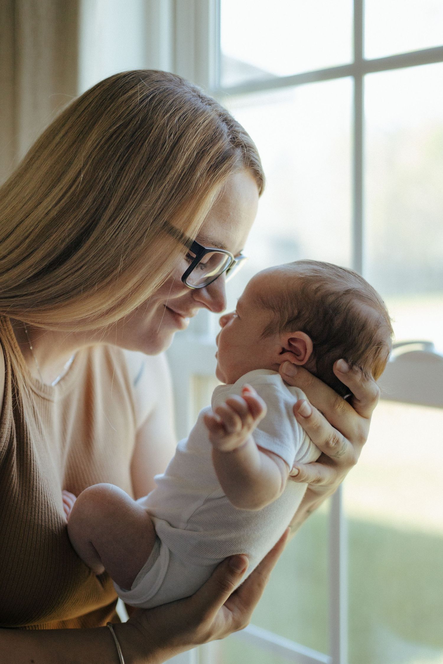 A tender moment between mother and baby by a sunlit window, sharing a gentle embrace in soft natural lighting.