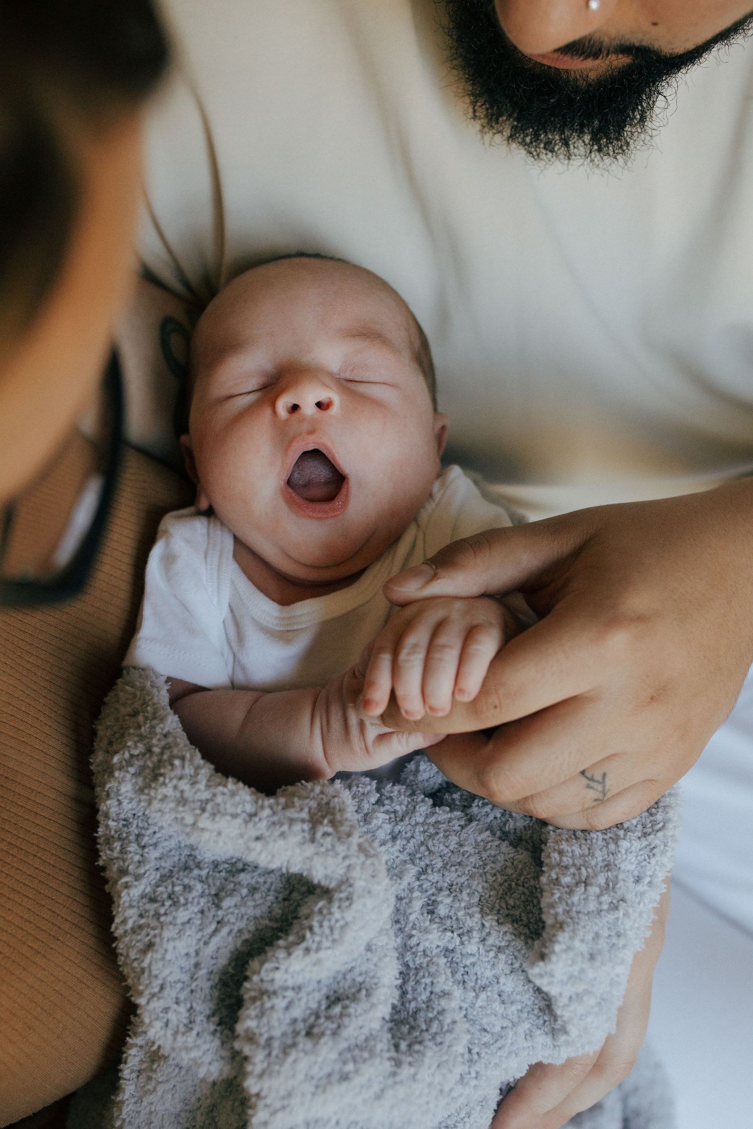 A sleepy baby yawns while wrapped in a cozy grey blanket, held by hands with a small tattoo visible.
