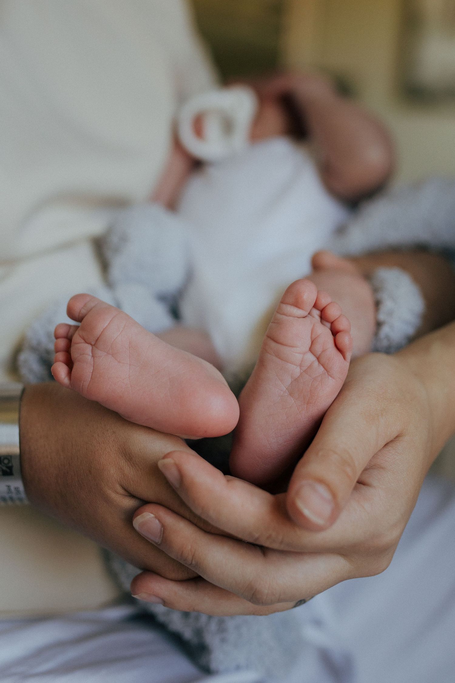 Tiny newborn baby feet gently cradled in caring hands against a soft white blanket and gray background.