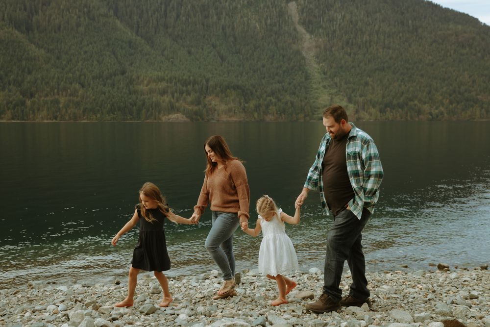A family holding hands while walking along a rocky lakeshore with mountains in the background.