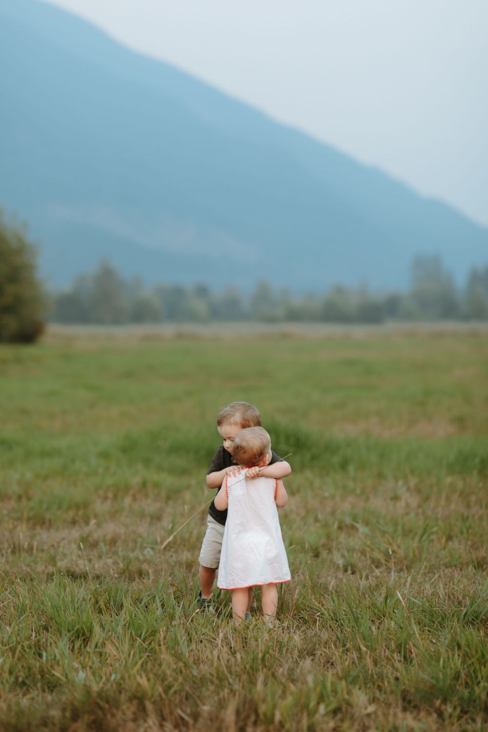 A small child in a white dress stands alone in a grassy field with mountains in the background.
