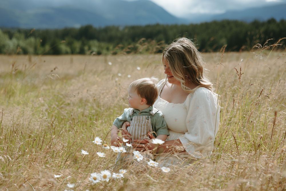 Parent and child sit together in a golden field with mountain views while dandelion fluff floats in the summer breeze.