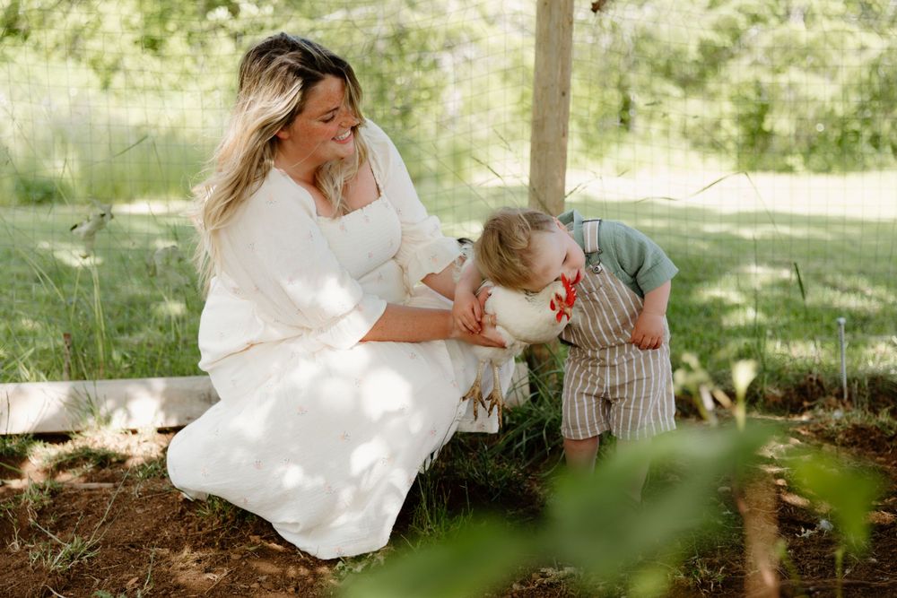A bride in a white dress sits in dappled sunlight on a rustic bench with a toddler on a sunny afternoon.