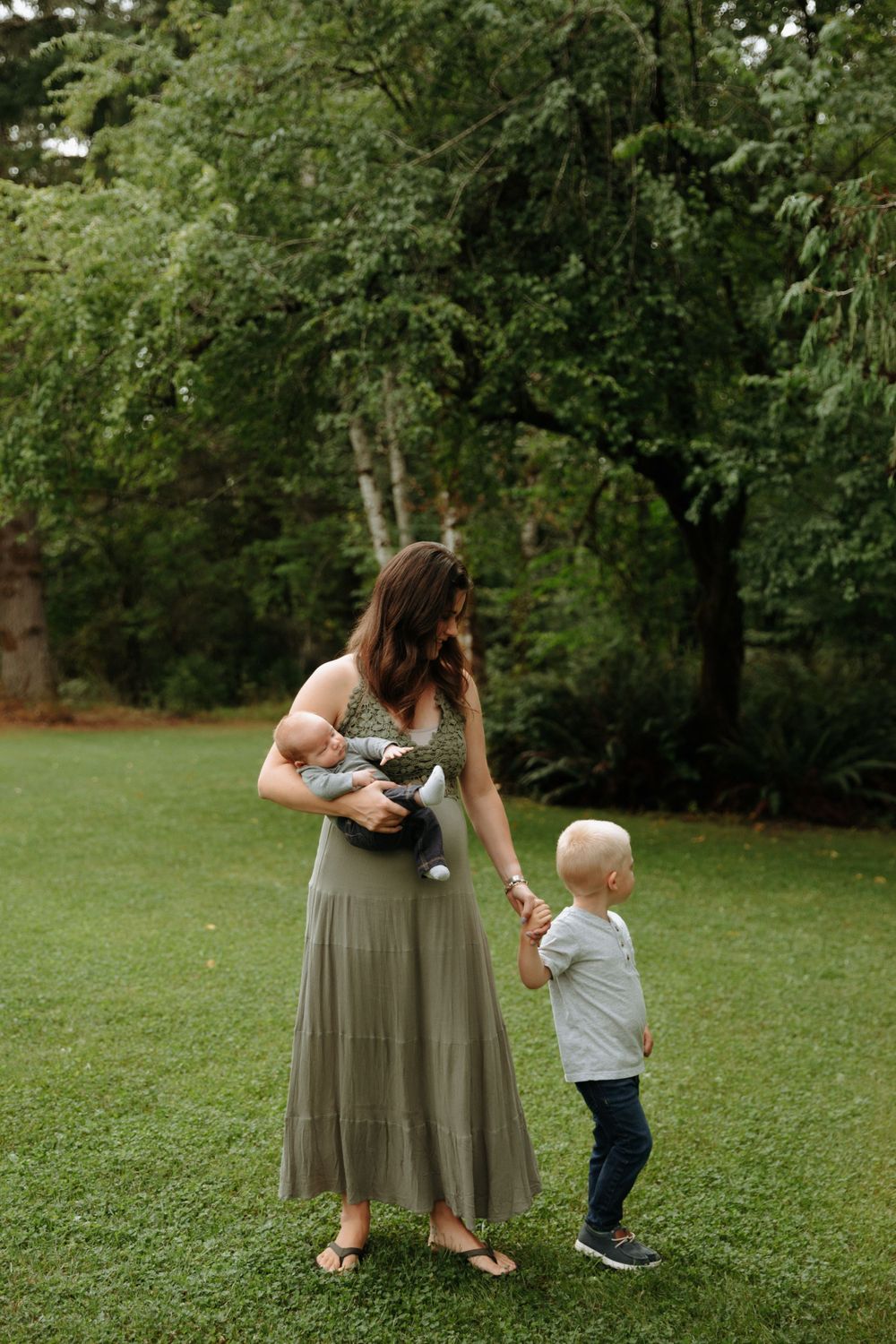 A woman in a flowing maxi skirt holds a baby carrier while a toddler stands beside her in a green outdoor setting.