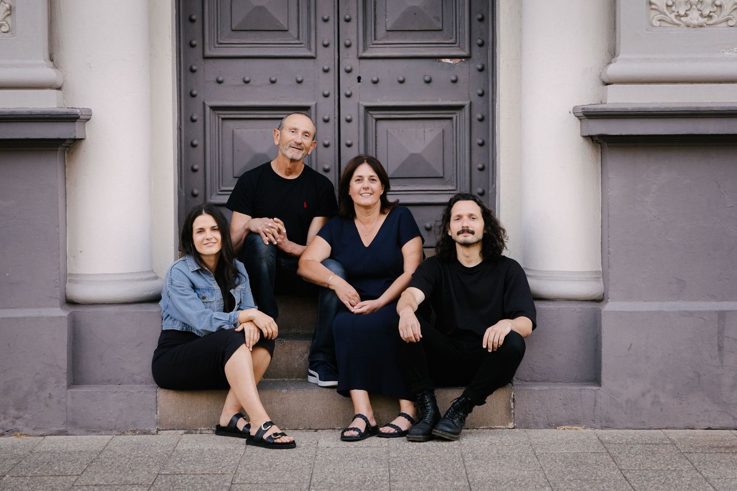 Group of four people sitting together on stone steps in front of ornate grey door.