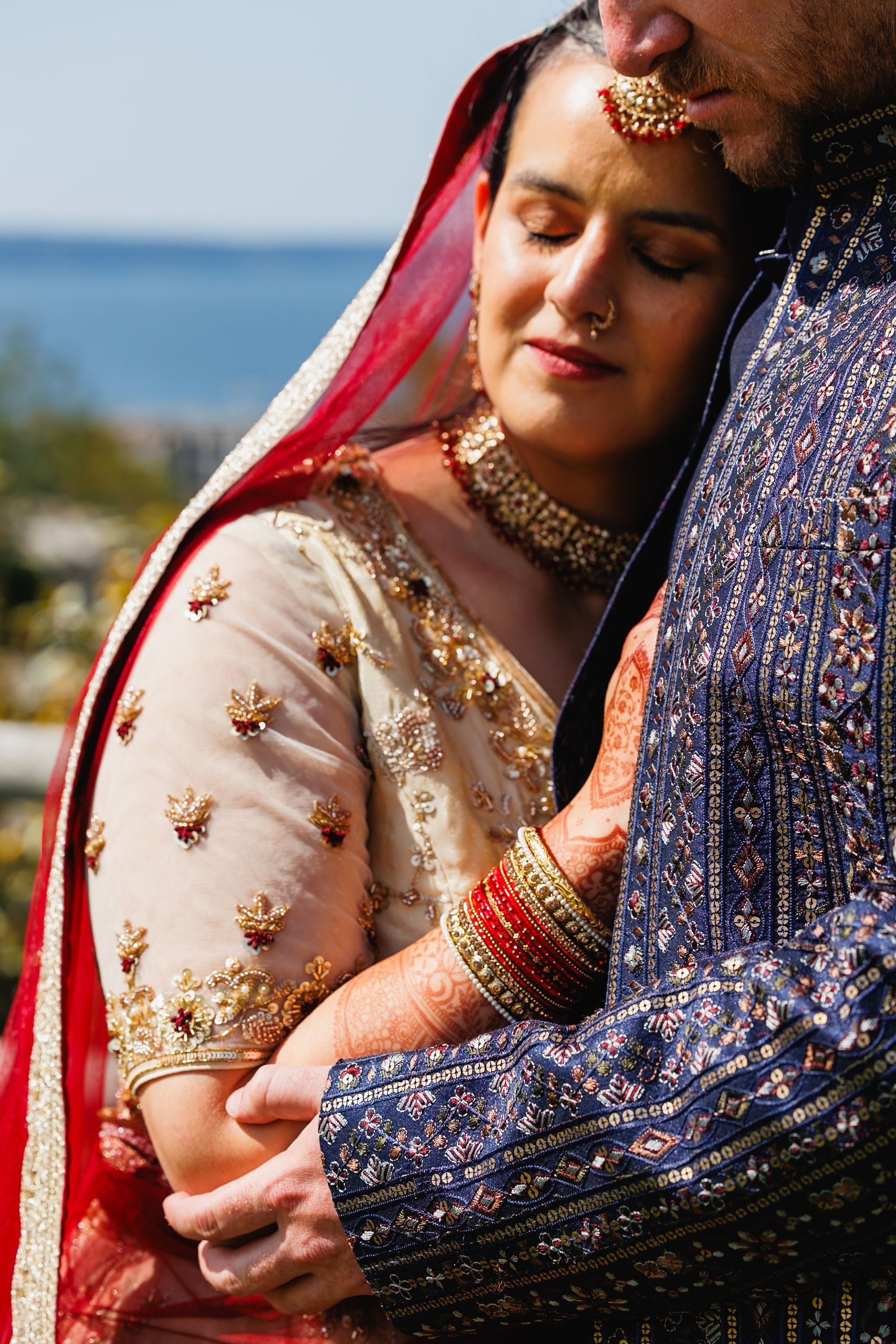 Bride in traditional red and gold Indian bridal attire embraces groom in blue embroidered sherwani.