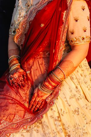 Close-up of Indian bride's hands adorned with intricate mehndi henna, bangles, and red dupatta.