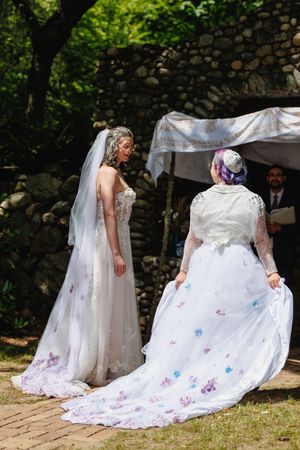 Two brides face each other under a chuppah during an outdoor Jewish wedding ceremony in a garden.