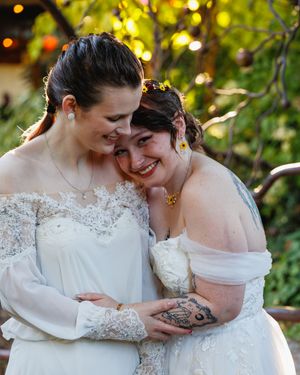 Two brides in white off-shoulder gowns share a joyful embrace outdoors at their wedding.