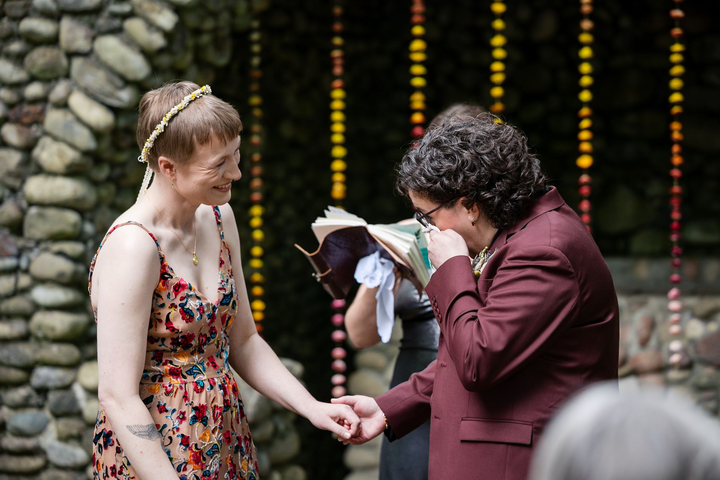 Two brides exchange rings during an emotional outdoor wedding ceremony with colorful garland backdrop.