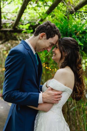 Smiling bride in off-shoulder white gown and groom in navy suit share tender moment in lush garden setting.