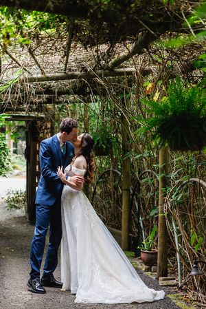 Bride and groom share a kiss under rustic wooden pergola surrounded by ferns and greenery in garden venue.