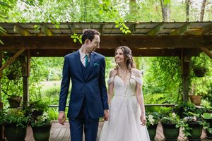 Happy newlyweds walk hand-in-hand on wooden deck beneath pergola in beautiful green garden wedding venue.
