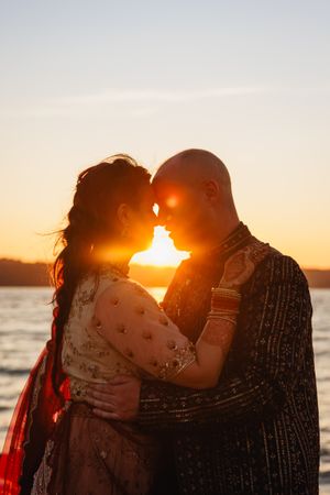 Indian couple in traditional wedding attire share romantic sunset moment by the water, silhouetted against golden sky.