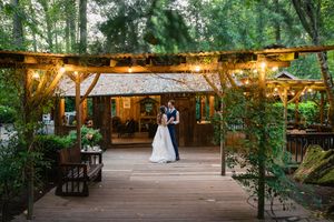 Newlyweds embrace under string lights on wooden deck at enchanting forest wedding venue at twilight.