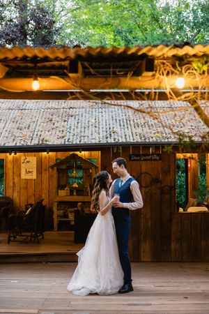 Bride and groom share first dance on wooden deck outside glowing rustic barn wedding venue at dusk.