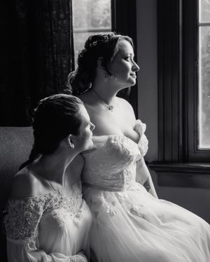 Two brides in white lace gowns pose elegantly by a window in a black and white wedding photo.