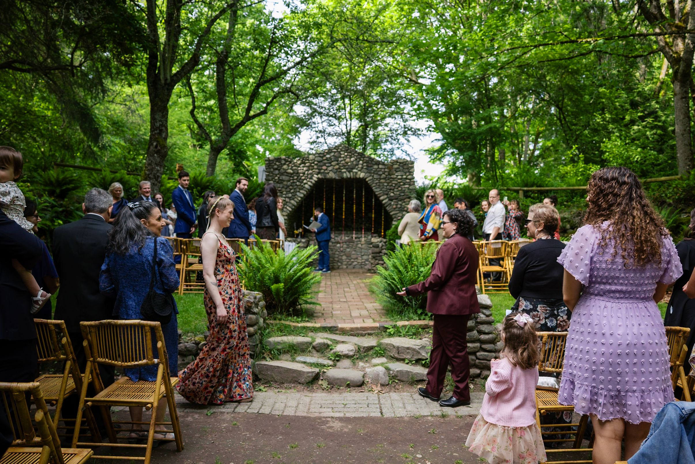 Outdoor wedding ceremony in a lush garden with guests seated on wooden chairs along a stone path.