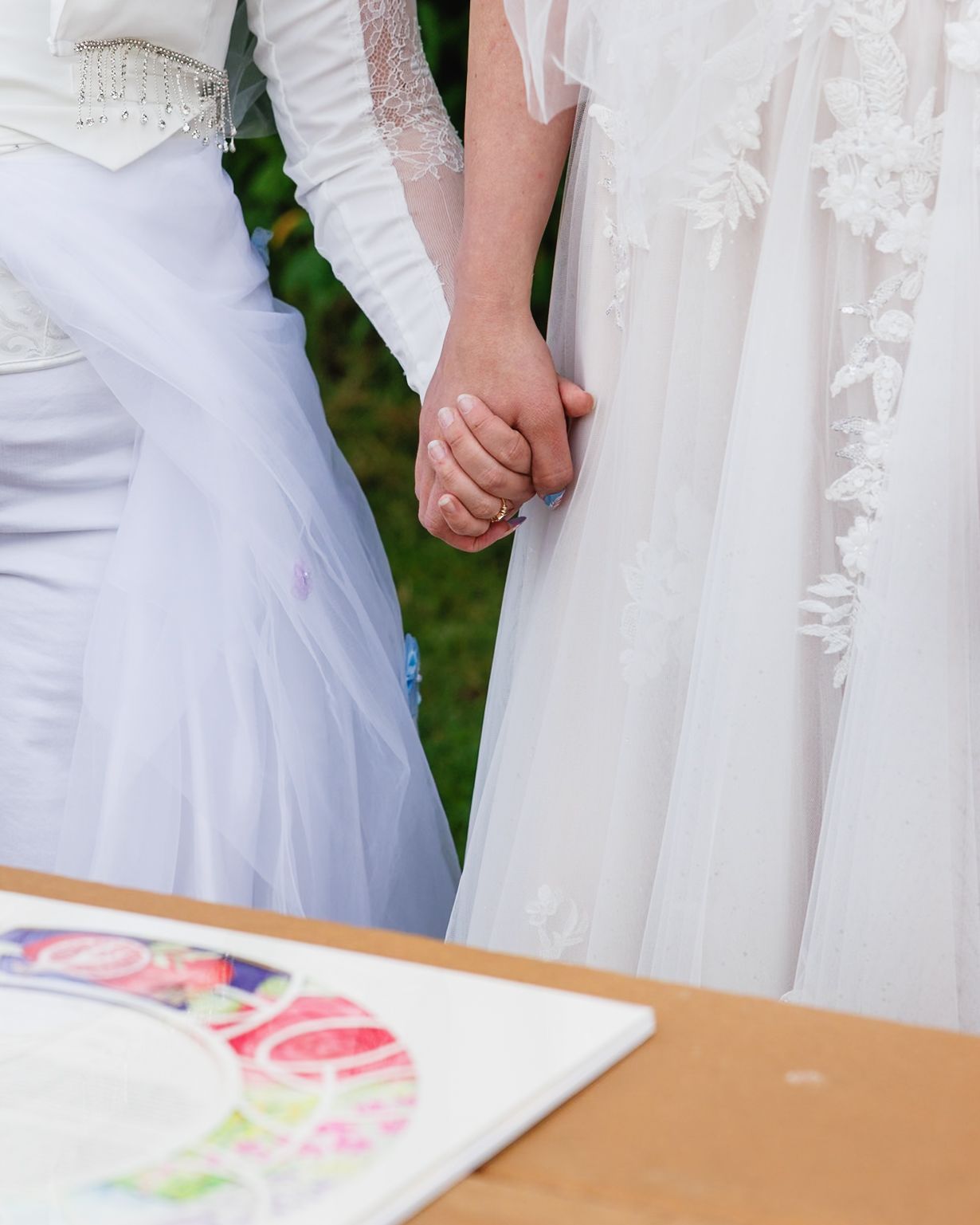 Two brides in white wedding gowns hold hands near a colorful ketubah signing table outdoors.