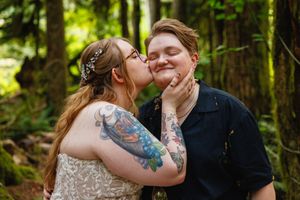 Tattooed bride kisses her partner's cheek in a lush forest wedding setting.