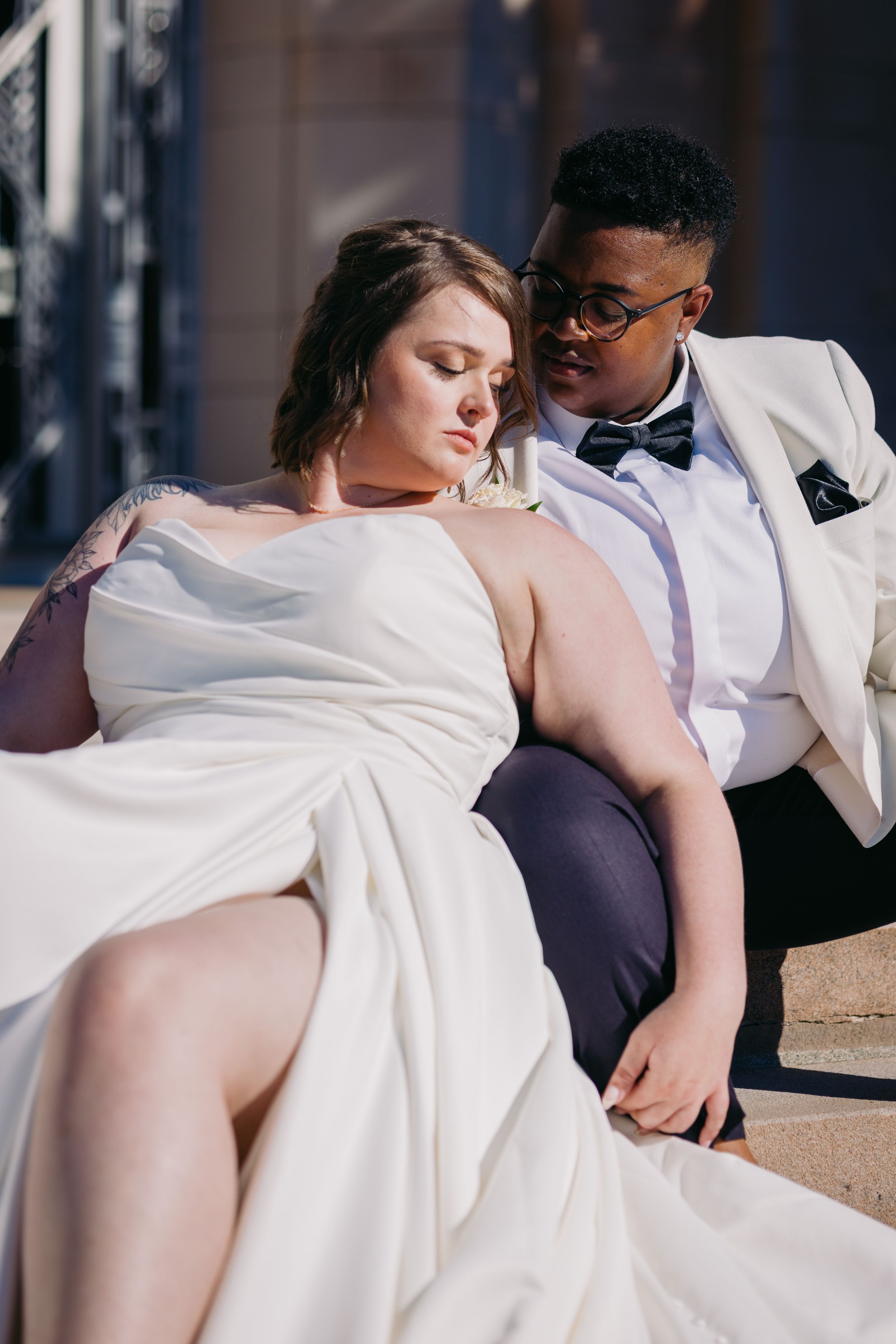 Interracial couple in wedding attire pose together on sunlit steps in a romantic portrait.