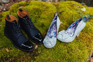 Black lace-up boots and sparkling silver heels with peacock feathers rest on a mossy rock.