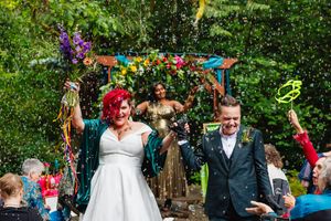 Newlywed couple celebrates with confetti shower under a colorful floral arch at outdoor wedding.