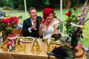 Newlywed couple toasts with pink drinks at a gold glitter sweetheart table with GAY marquee letters.