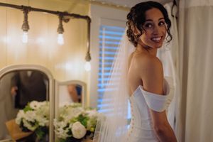 Smiling bride in an off-shoulder gown and veil poses in a warmly lit dressing room.