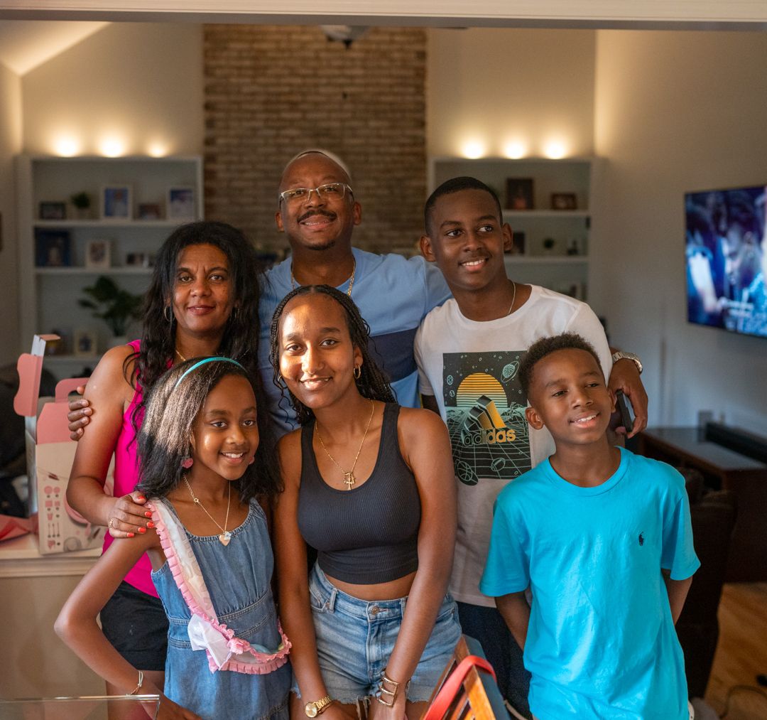 A happy family of five poses together in a cozy living room with exposed brick wall and warm lighting.