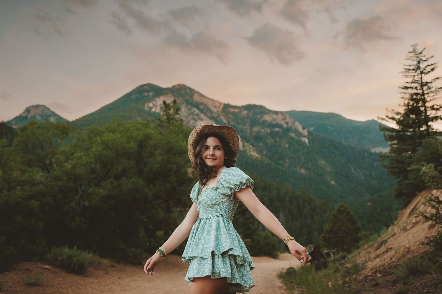 Someone in a mint dress and sun hat stands with arms outstretched on a mountain trail during golden hour.