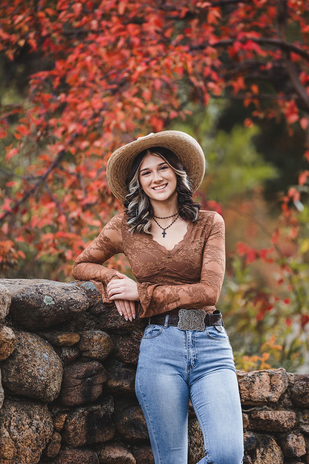Autumn portrait near stone wall with vibrant red maple leaves, featuring brown lace top, straw hat and blue jeans.