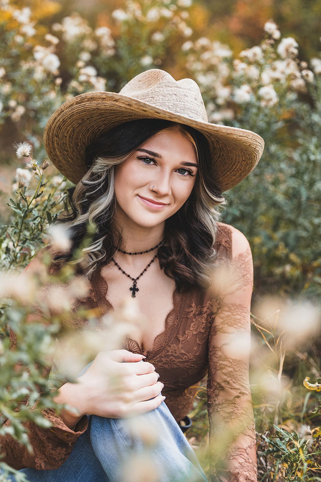 A rustic outdoor portrait featuring a straw hat wearing figure in a brown top amid blooming white flowers and greenery.