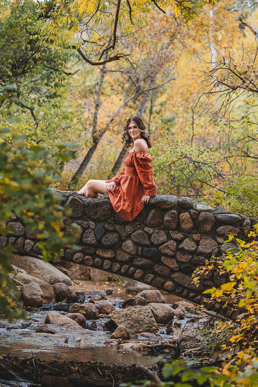 Someone in a burnt orange dress sits on a rustic stone bridge over a creek surrounded by autumn foliage in a park setting.