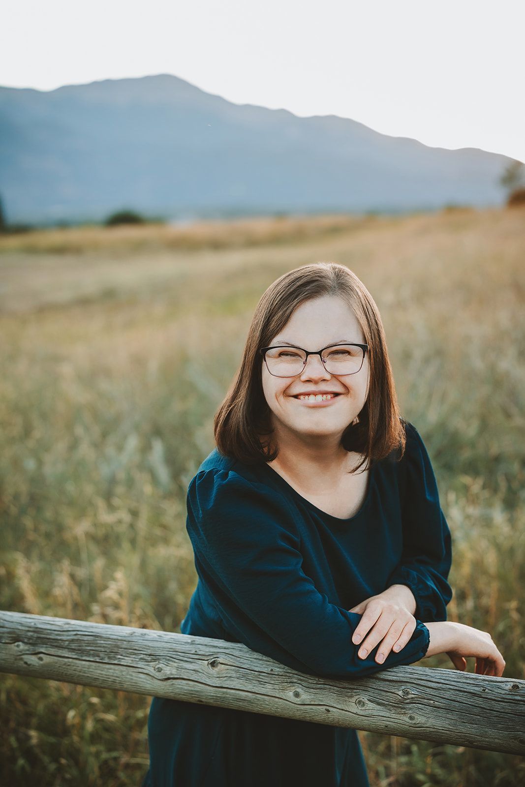High school senior  in glasses and navy sweater leans against wooden fence with mountains in background at golden hour.