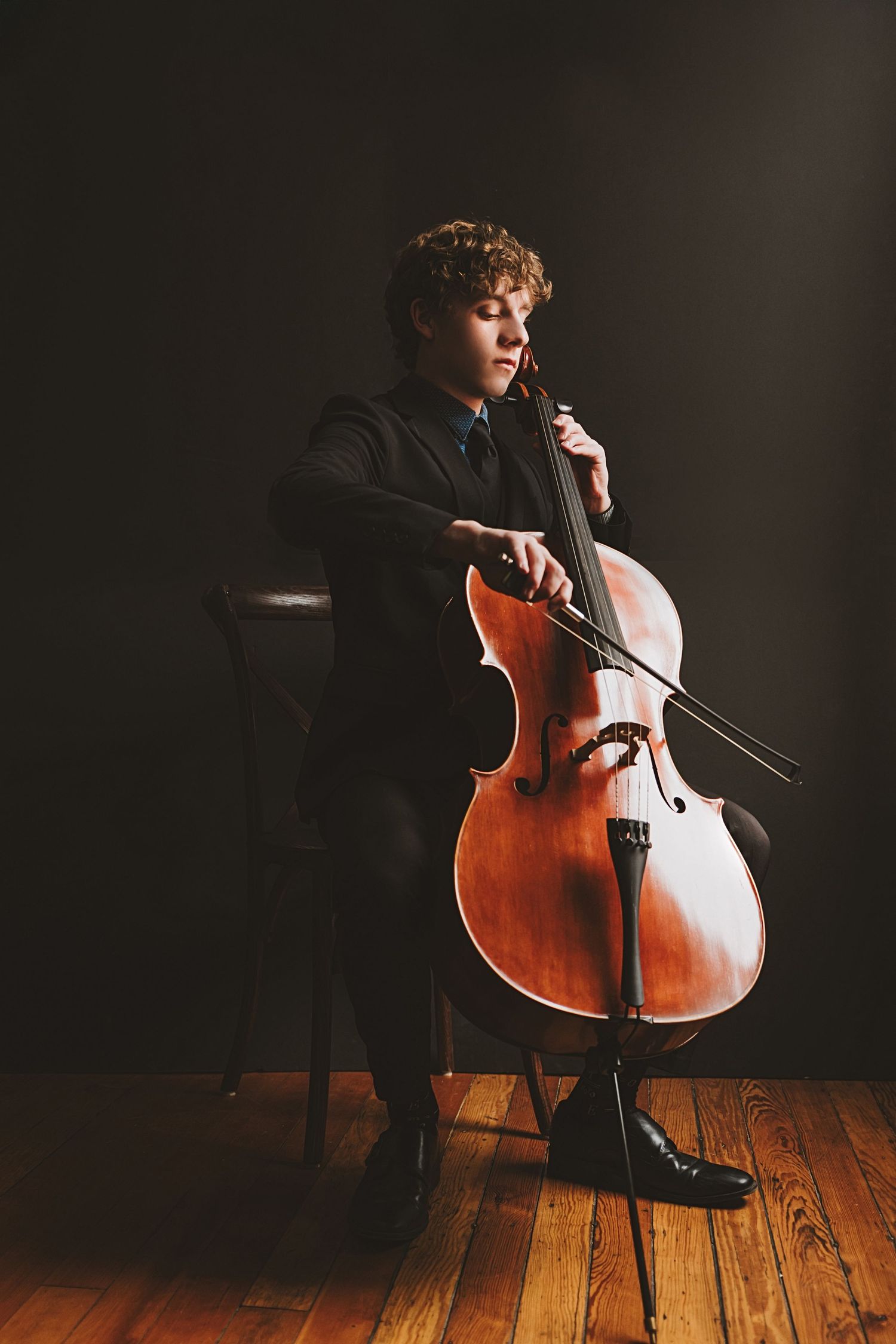 A musician in a black outfit performs on a wooden cello against a dark background on hardwood flooring.
