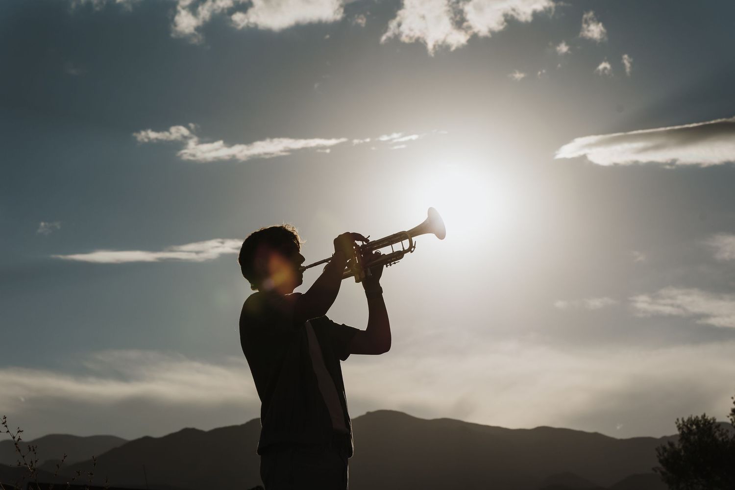 Silhouette of a trumpet player performing against a dramatic sunset sky with mountains in the background.